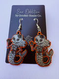 A pair of dangling skeleton mermaid earrings displayed on a black card that reads 'Ear Oddities by October House Co.' The earrings have bright orange edges, silver-toned metal hooks, and gray engraved mermaid and skull designs on a white background.