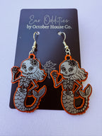 A pair of dangling skeleton mermaid earrings displayed on a black card that reads 'Ear Oddities by October House Co.' The earrings have bright orange edges, silver-toned metal hooks, and gray engraved mermaid and skull designs on a white background.