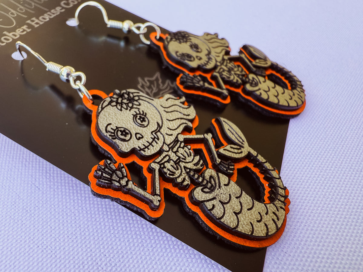 A pair of dangling skeleton mermaid earrings with silver fishhook wires and a black, gray, and bright orange layered design, displayed on a black branded card over a light lavender textured background.
