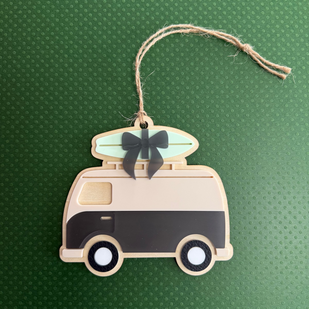 A wooden camper-shaped ornament with a natural wood finish, featuring a black and white lower section, a light green surfboard on top, a black bow, and a twine hanger, displayed on a textured green background.