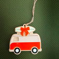 A retro-style wooden van ornament with a surfboard on top, tied with a red bow, hanging against a dark green textured background.