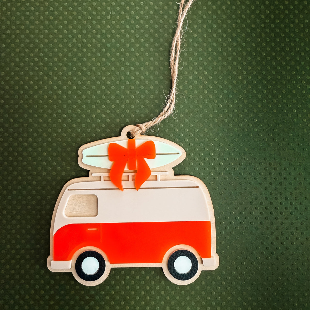 A retro-style wooden van ornament with a surfboard on top, tied with a red bow, hanging against a dark green textured background.
