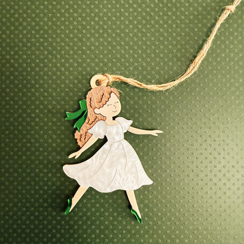 A handcrafted wooden fairy ornament with a white dress, green shoes, and brown hair, hanging from a natural jute string against a textured green background.
