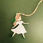 A handcrafted wooden fairy ornament with a white dress, green shoes, and brown hair, hanging from a natural jute string against a textured green background.