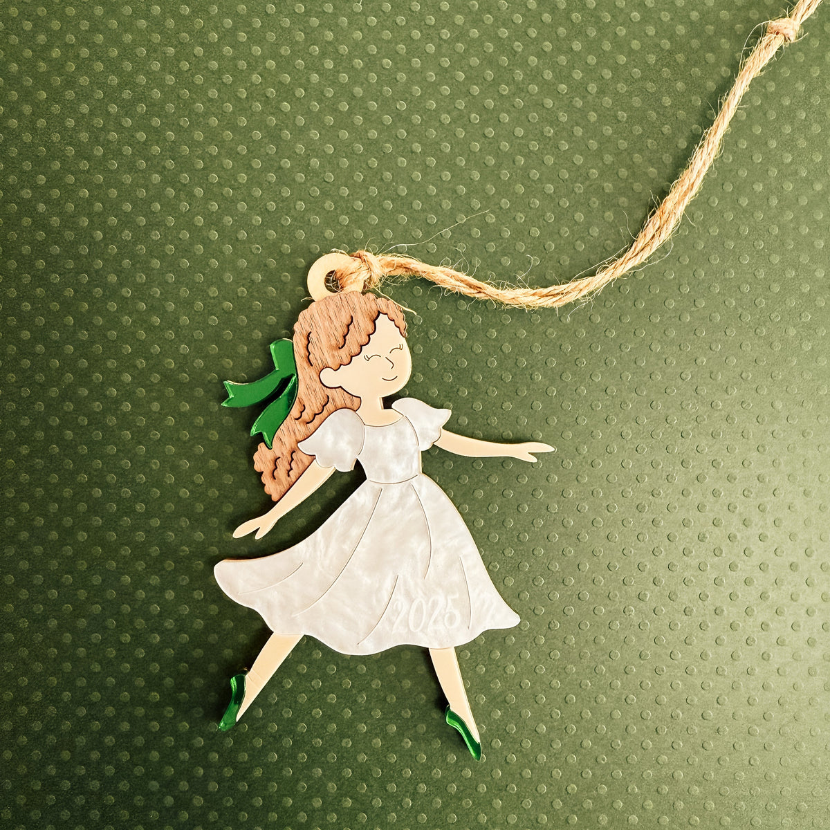A handcrafted wooden fairy ornament with a white dress, green shoes, and brown hair, hanging from a natural jute string against a textured green background.
