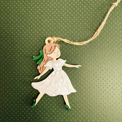A handcrafted wooden fairy ornament with a white dress, green shoes, and brown hair, hanging from a natural jute string against a textured green background.