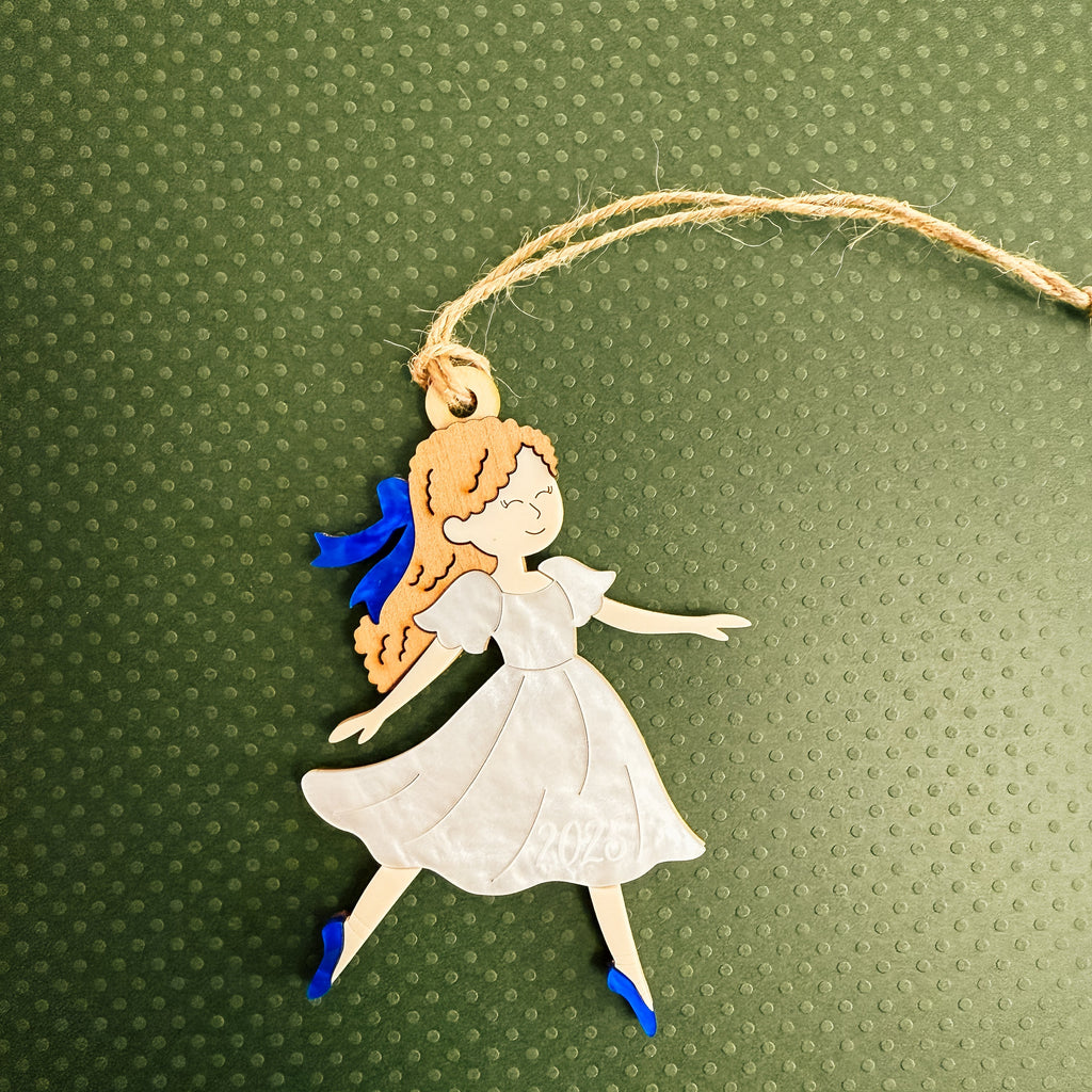 A die-cut hanging ornament featuring a cartoon girl with orange hair, a white dress, and blue shoes, suspended by a twine string against a textured dark green background.