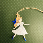 A die-cut hanging ornament featuring a cartoon girl with orange hair, a white dress, and blue shoes, suspended by a twine string against a textured dark green background.