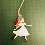 A decorative hanging ornament shaped like a girl in a white dress with brown hair tied back with a red ribbon, red shoes, and a natural jute hanging string, shown on a textured dark green background.