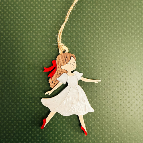 A decorative hanging ornament shaped like a girl in a white dress with brown hair tied back with a red ribbon, red shoes, and a natural jute hanging string, shown on a textured dark green background.