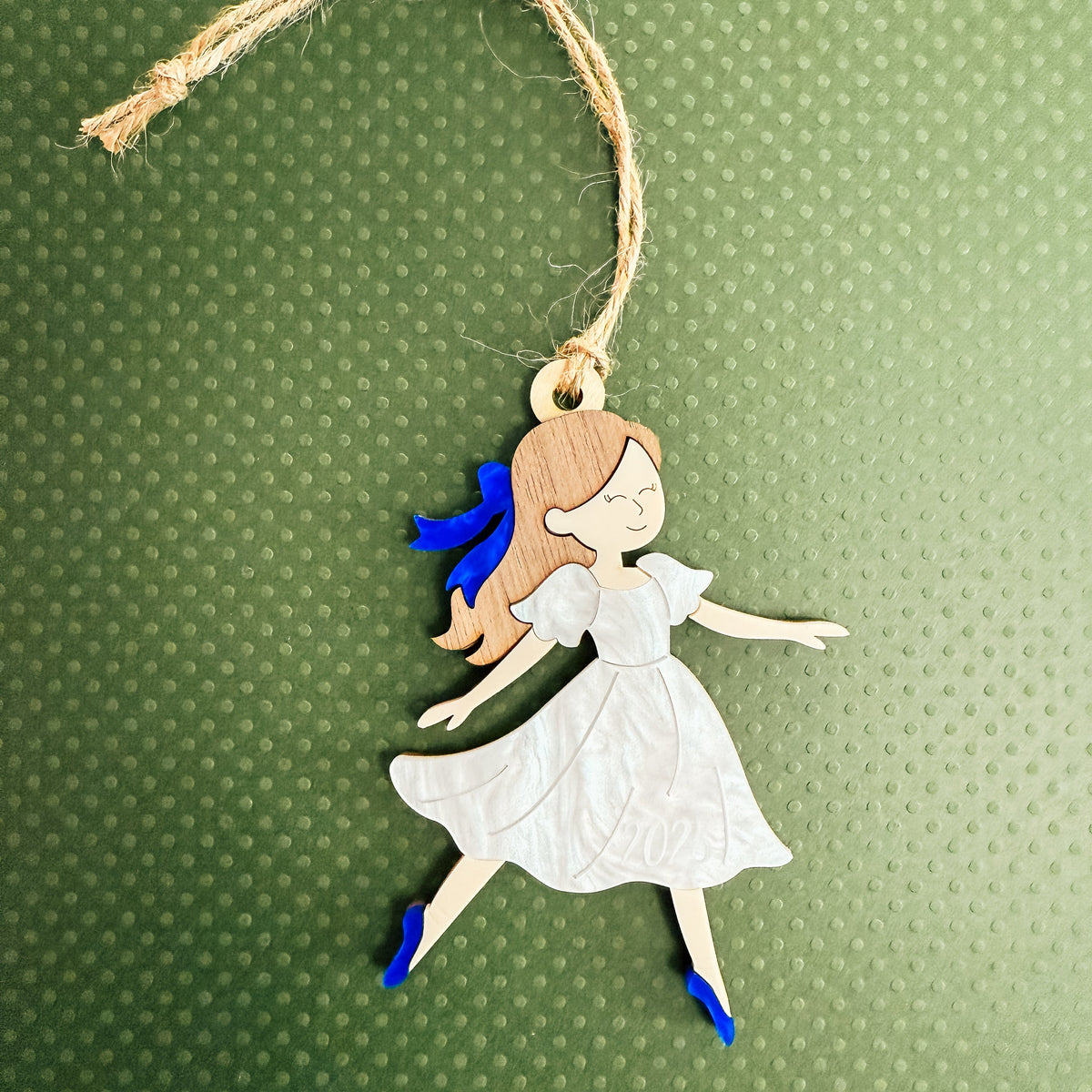 A handcrafted wooden ornament shaped like a dancing girl in a white dress with blue shoes and a blue bow, hanging from a jute twine loop against a textured green background.