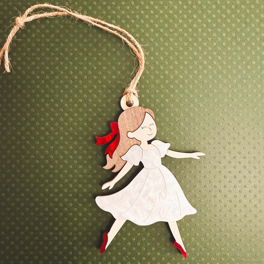 A handcrafted wooden ornament shaped like a dancing girl in a white dress with red shoes and a red bow, hanging from a jute twine string against a textured olive green background.