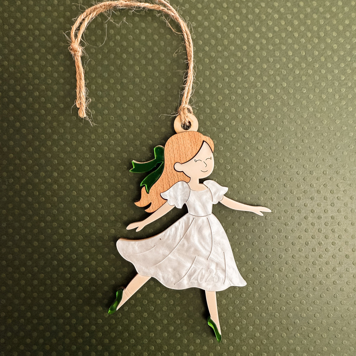 A handcrafted wooden fairy ornament featuring a white dress, orange hair, and green accents, hanging from a natural jute string against a textured olive green background.
