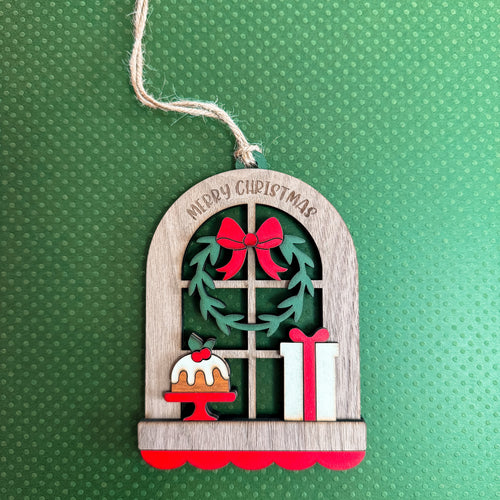 Arched wood textured ornament with "Merry Christmas" engraved featuring a window display of a wreath, cake, and present. On a green textured background.