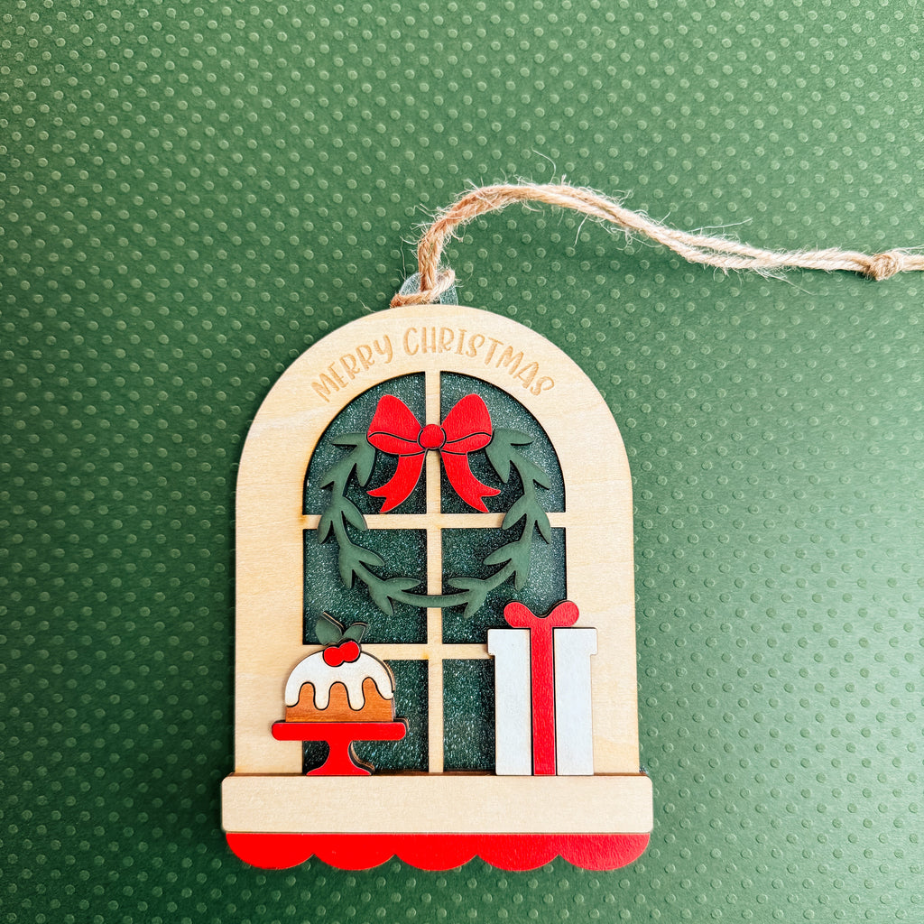 A wooden Christmas window ornament with a hanging string, featuring a green wreath with red bows, a small cake, a gift box, and 'MERRY CHRISTMAS' written on the arched top, set against a textured green background.