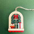 A wooden Christmas window ornament with a hanging string, featuring a green wreath with red bows, a small cake, a gift box, and 'MERRY CHRISTMAS' written on the arched top, set against a textured green background.