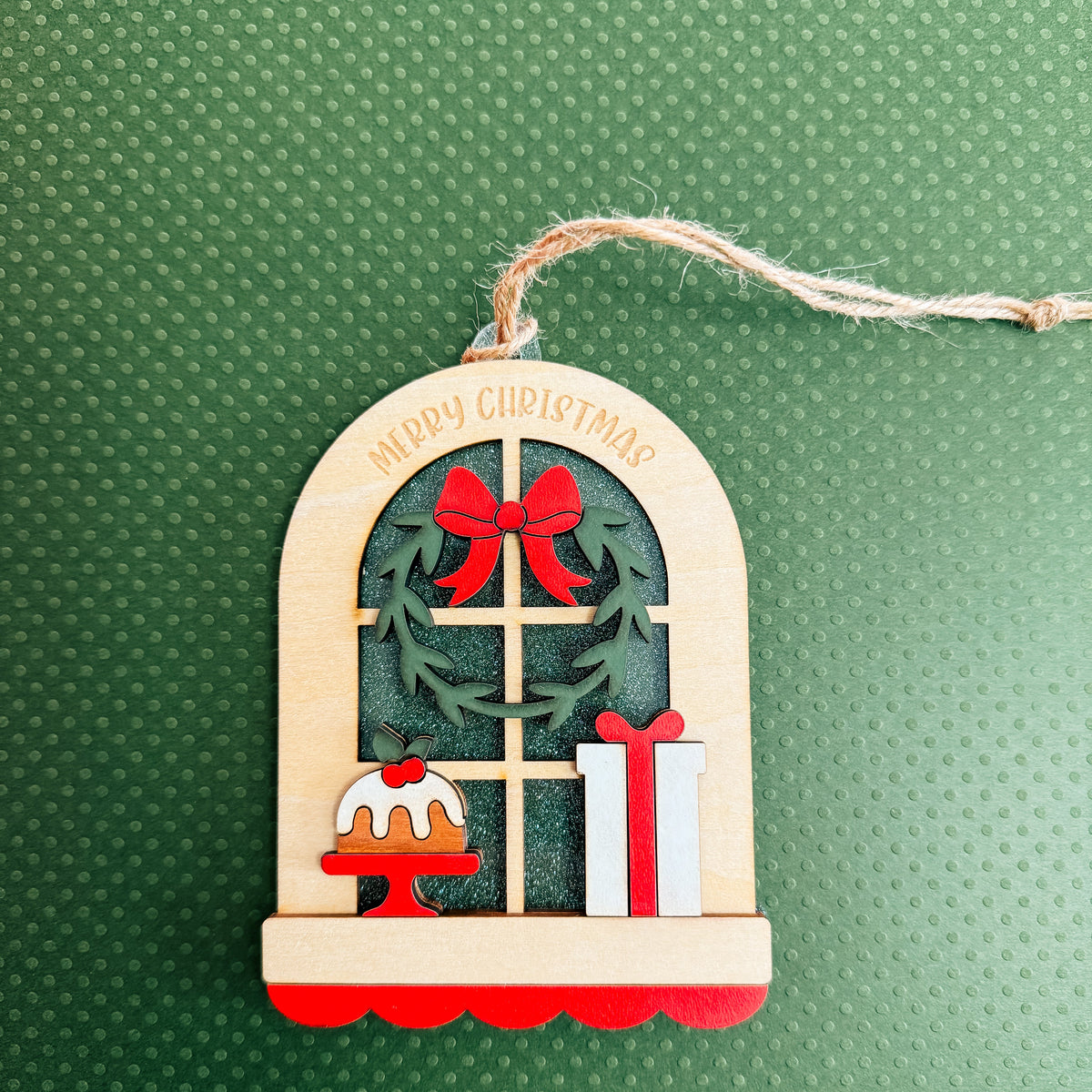 A wooden Christmas window ornament with a hanging string, featuring a green wreath with red bows, a small cake, a gift box, and 'MERRY CHRISTMAS' written on the arched top, set against a textured green background.
