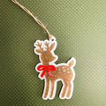 A die-cut Christmas ornament shaped like a brown deer with white spots, a red bow, and a jute hanging string, shown on a textured olive green background.