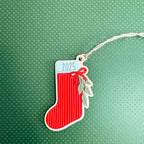 A red Christmas stocking ornament with white vertical stripes, a white top label reading '2025', a red bow, and green leaf accents, hanging from a white string on a textured green background.