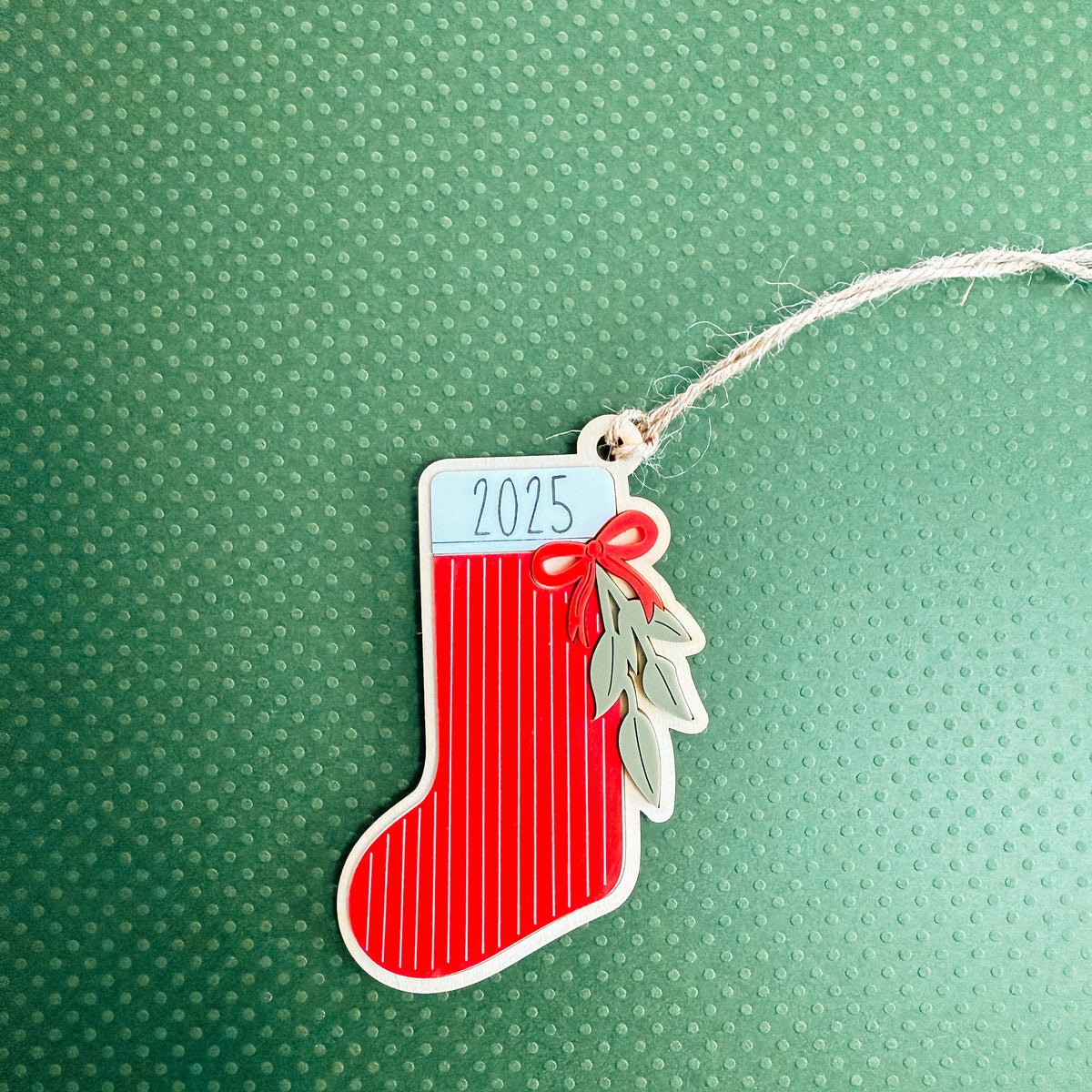 A red Christmas stocking ornament with white vertical stripes, a white top label reading '2025', a red bow, and green leaf accents, hanging from a white string on a textured green background.