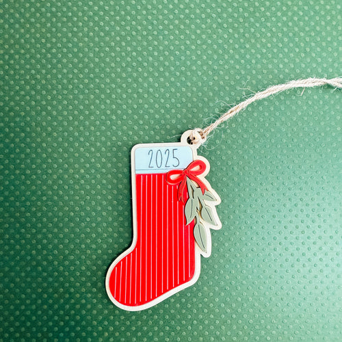 A red Christmas stocking ornament with white vertical stripes, a white top label reading '2025', a red bow, and green leaf accents, hanging from a white string on a textured green background.