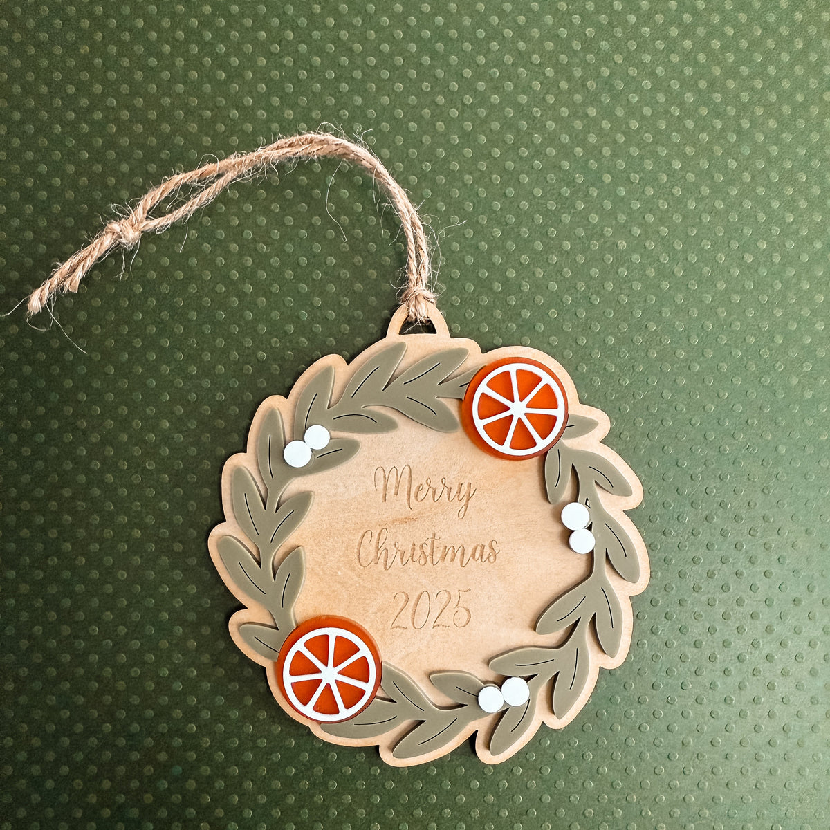 A personalized wooden Christmas ornament shaped like a wreath, hanging by a twine loop. The ornament is light natural wood with dark brown engraved leaf details, featuring two orange slice decorations and small white bead accents.