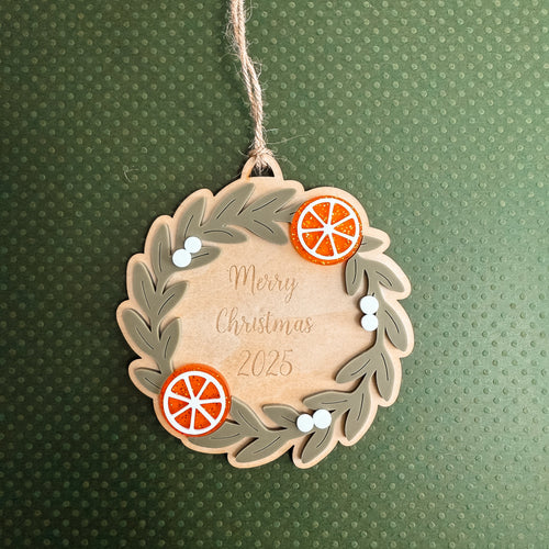 A wooden Christmas ornament shaped like a wreath, hanging by a twine string. The ornament features a light wood center with the words 'Merry Christmas 2025' in a cursive font, surrounded by layered gray-green leafy wreath edges and three orange citrus slice decorations with small white berry accents.