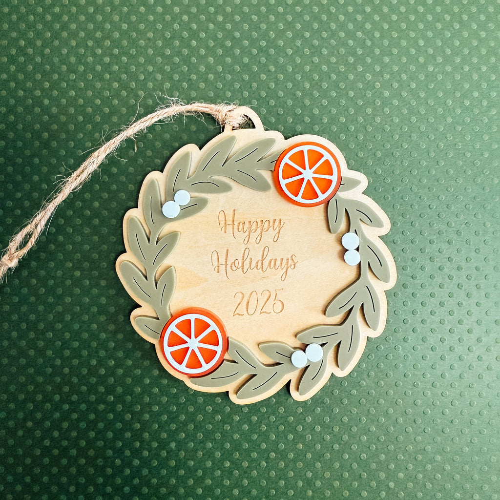 A personalized wooden Christmas ornament shaped like a wreath, featuring the text 'Happy Holidays 2025' in gold foil, decorated with olive green leaves, orange slice accents, and small blue berries. The ornament hangs from a rustic twine string against a textured dark green background.