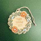 A round wooden hanging ornament with a wreath design, featuring the text 'Happy Holidays 2025' in gold script. The ornament is decorated with two orange slice accents, small white berry embellishments, and a natural jute hanging string, set against a textured dark green background.