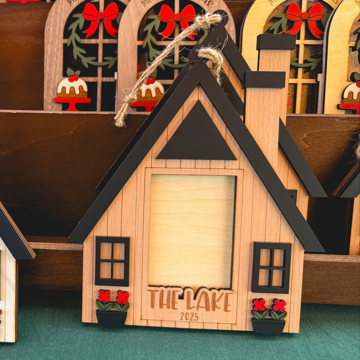 A set of small house-shaped wooden picture frames with black-trimmed roofs, window details, red flower accents, and blank beige photo inserts, displayed on a green surface with other decorative items in the background.