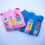 Two folded sweatshirts, one pink and one blue, each featuring a colorful embroidered nutcracker character on the front, set against a light blue background.