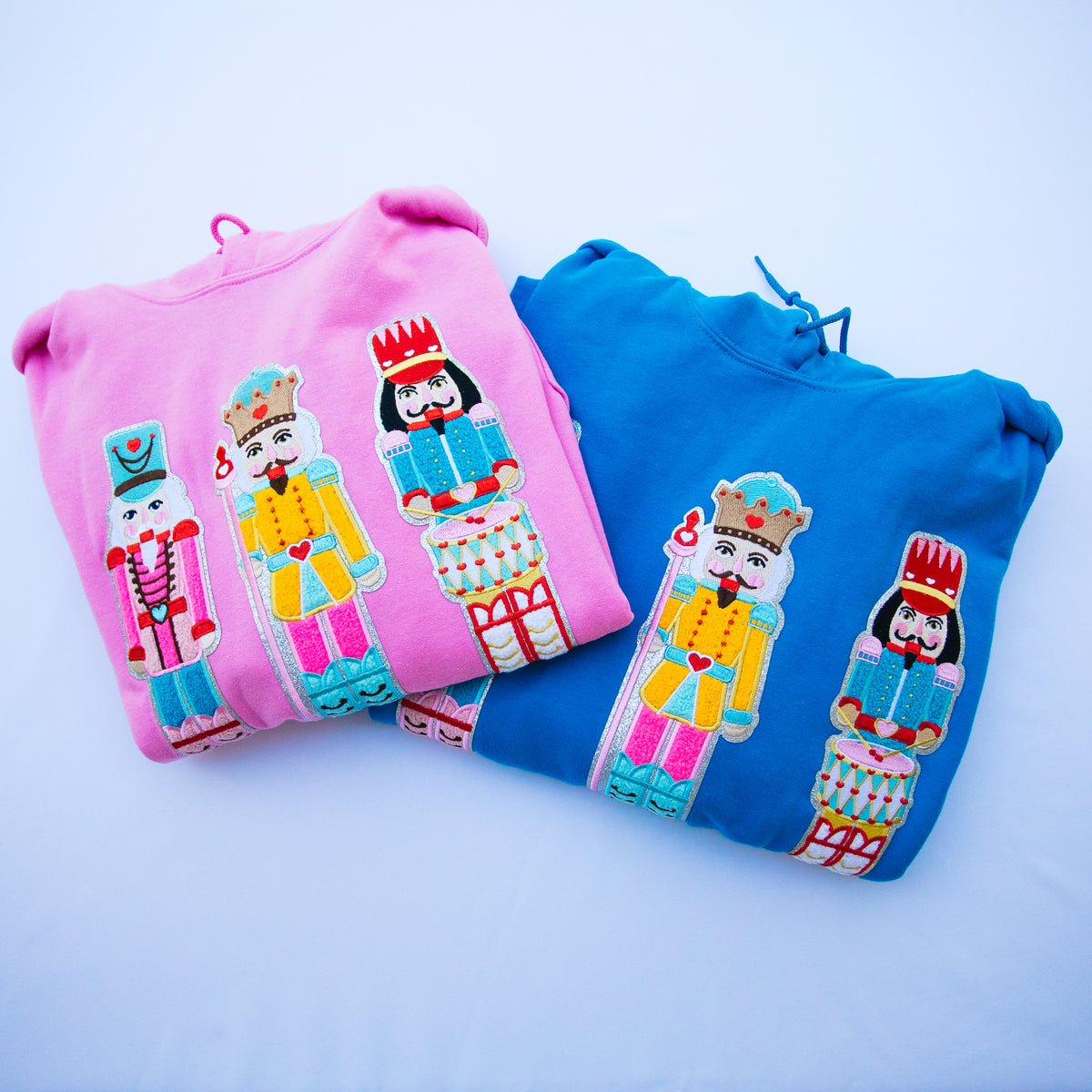 Two folded sweatshirts, one pink and one blue, each featuring a colorful embroidered nutcracker character on the front, set against a light blue background.