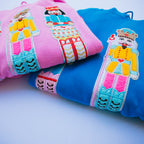 Two folded sweatshirts, one pink and one blue, stacked on a light background. Both feature a large embroidered nutcracker character on the front in yellow, pink, teal, red, and white.