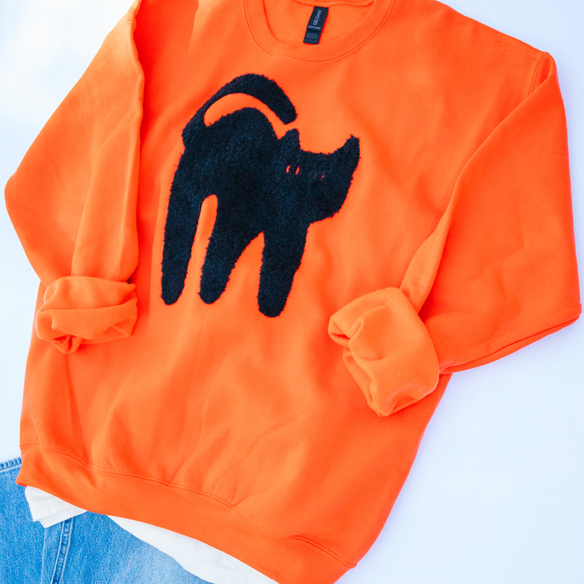 Bright orange crewneck sweatshirt featuring a large black cat silhouette applique on the front, styled flat with rolled cuffs and a bit of blue jeans visible at the bottom left.