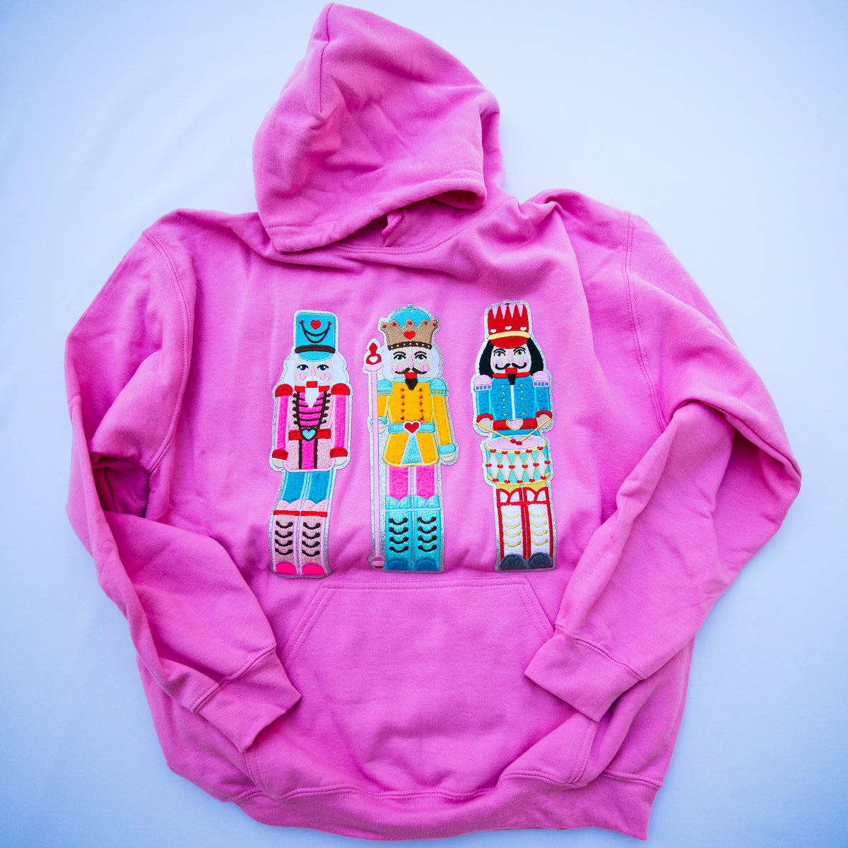 A bright pink pullover hoodie laid flat on a white background, featuring three colorful embroidered nutcracker figures across the chest.