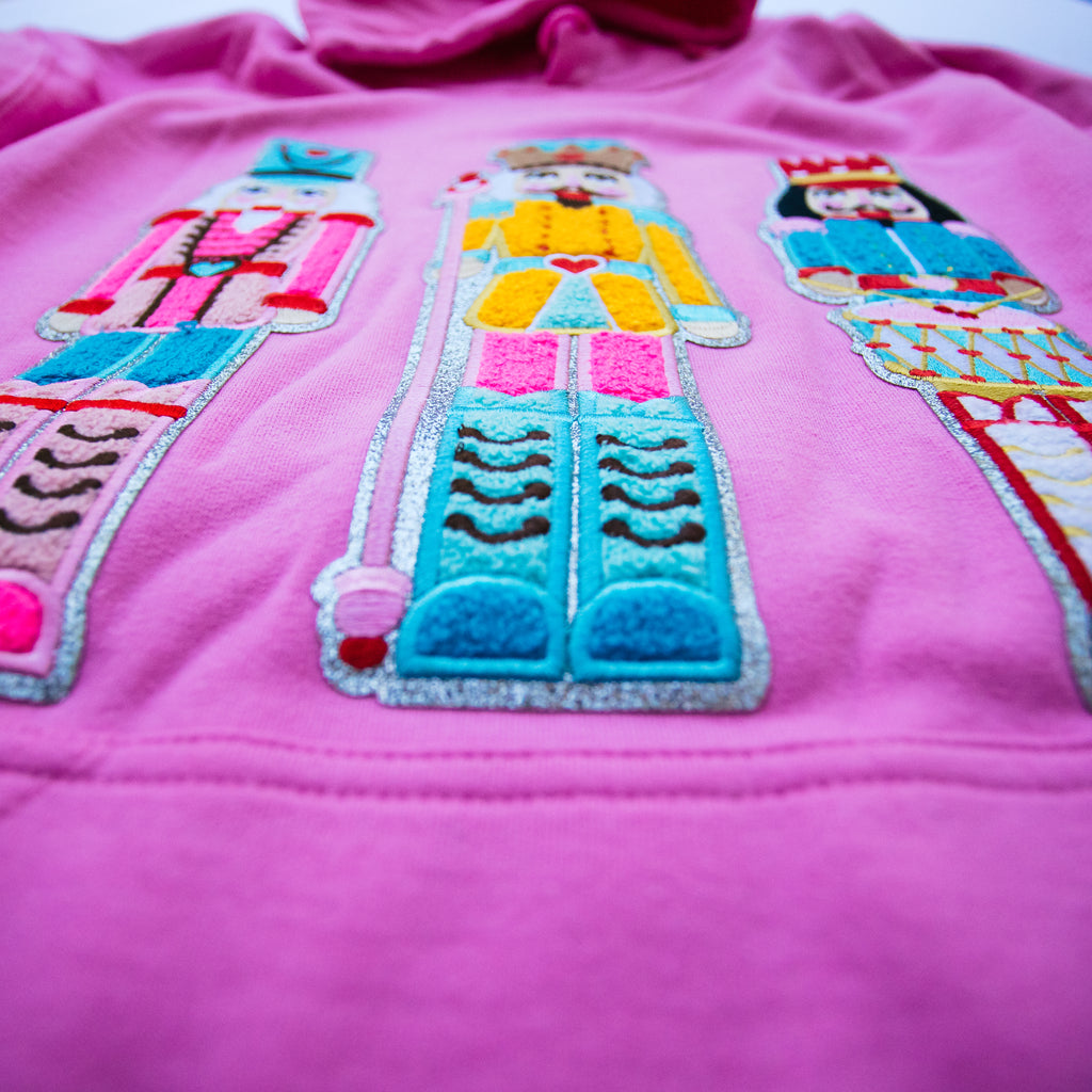 A close-up of a bright pink garment featuring three large, colorful embroidered nutcracker figures across the chest.