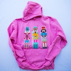 A bright pink hoodie laid flat on a light blue background, featuring three colorful embroidered nutcracker figures across the chest.