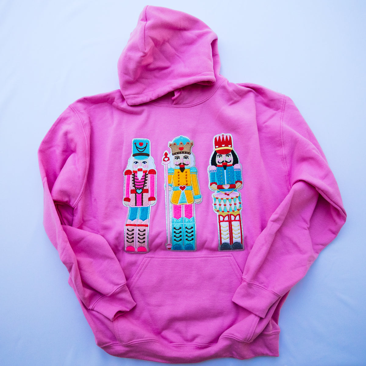A bright pink hoodie laid flat on a light blue background, featuring three colorful embroidered nutcracker figures across the chest.
