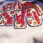 A close-up of a decorative embroidered patch featuring the word "GAM" in large red letters with leopard-print centers, outlined in white and glittery trim, on a light fabric background.