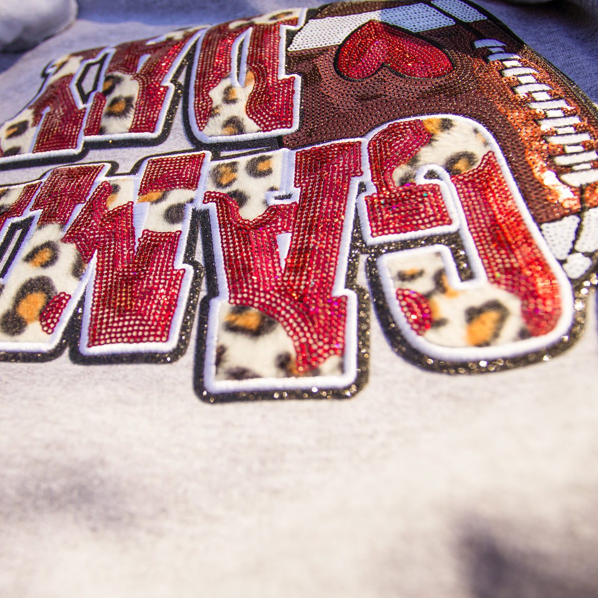 A close-up of a decorative embroidered patch featuring the word "GAM" in large red letters with leopard-print centers, outlined in white and glittery trim, on a light fabric background.