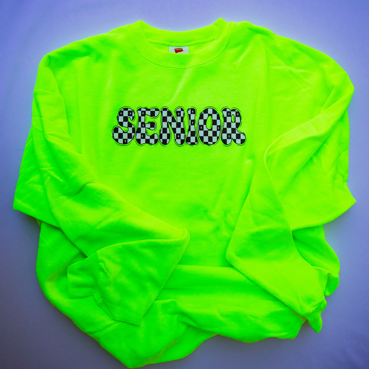A neon green long-sleeve sweatshirt with 'SENIOR' printed across the chest in a black-and-white checkered pattern, laid flat on a purple background.