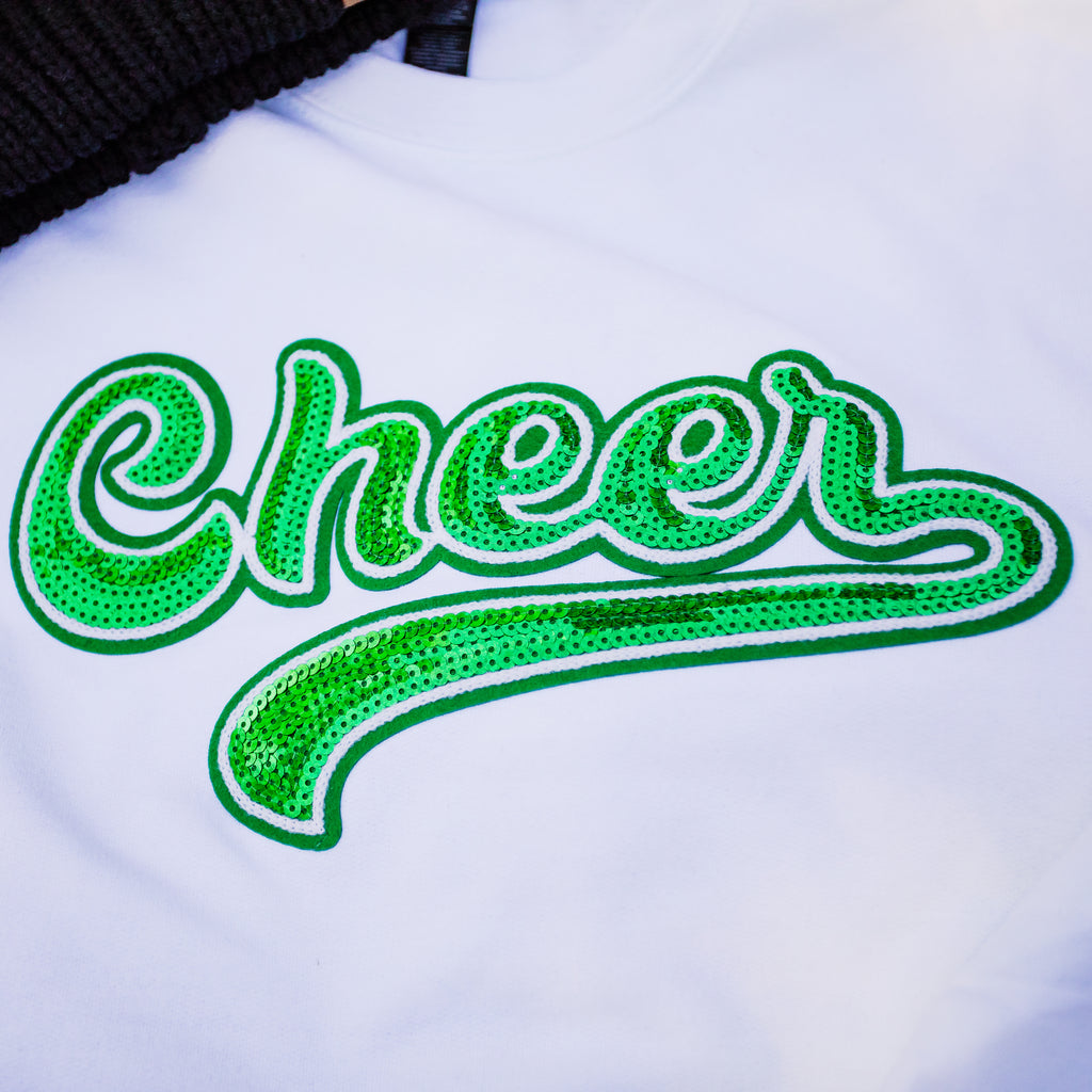 A close-up of a white t-shirt featuring a large, cursive 'Cheer' graphic embellished with green sequins and a white outline.