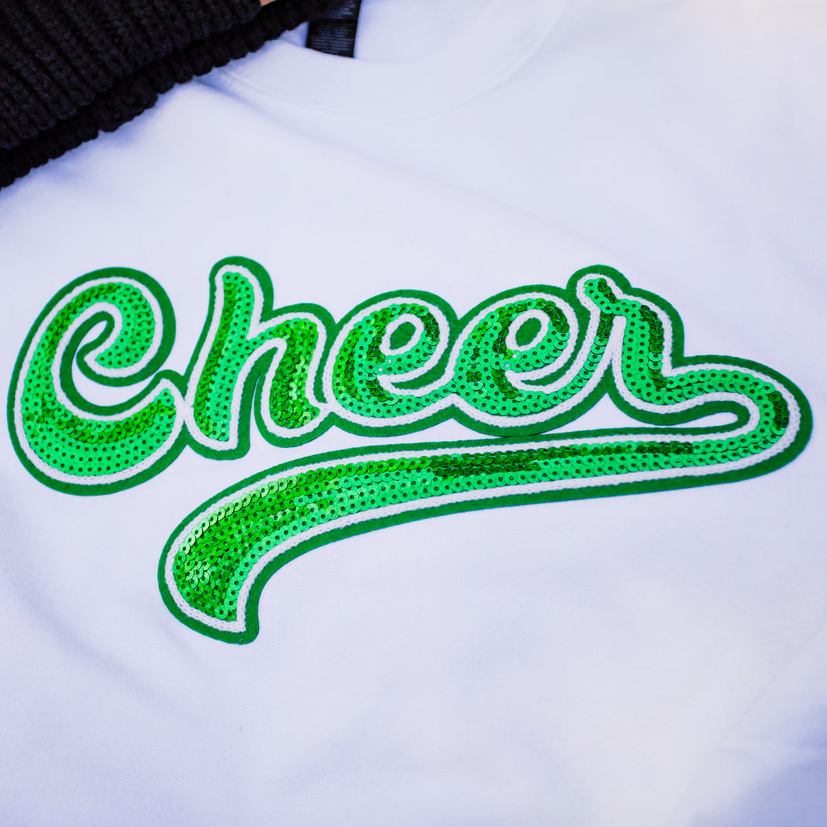 A close-up of a white t-shirt featuring a large, cursive 'Cheer' graphic embellished with green sequins and a white outline.