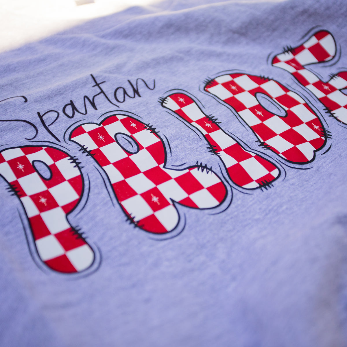 A close-up of a light lavender heather t-shirt featuring a graphic design. The design displays the word 'PRIDE' in large, stylized block letters filled with a red and white checkered pattern, outlined in black with small star details, and the word 'Spartan' written in black cursive script above it.