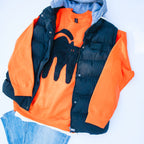 A styled flat-lay of a children's hooded puffer vest layered over an orange sweatshirt with a black cat graphic, paired with blue jeans on a white background.