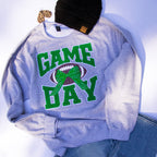 A light heather gray crewneck sweatshirt with a 'GAME DAY' football graphic in green and brown, styled flat-lay with a black knit beanie and blue jeans on a white background.