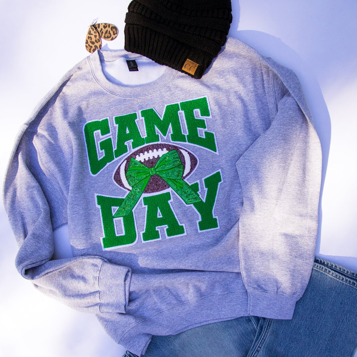 A light heather gray crewneck sweatshirt with a 'GAME DAY' football graphic in green and brown, styled flat-lay with a black knit beanie and blue jeans on a white background.
