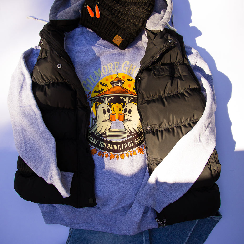 A styled flat-lay of a light blue graphic sweatshirt layered under a black puffer vest with light gray lining, accessorized with a black knit beanie and blue jeans against a white background.