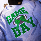 A close-up of a heather gray sweatshirt featuring a 'GAME DAY' graphic with a football and green bow. A black knit beanie with a tan logo patch is partially visible at the top.