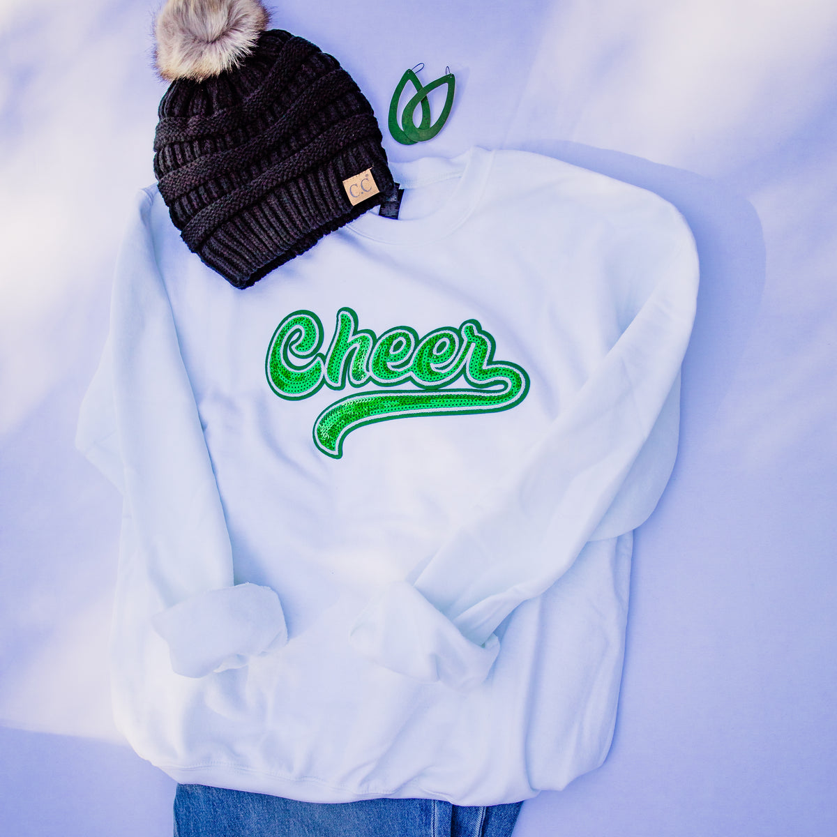 A light blue sweatshirt with green embroidered 'Cheer' lettering, styled in a flat lay with a black knit beanie featuring a beige faux-fur pom-pom and a small green embroidered leaf patch.